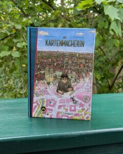 Die Kartenmacherin - die Graphic Novel steht auf einer dunkelgrünen Balkonbrüstung.