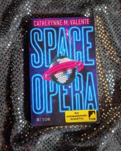 Space Opera - der metaglaktische Grand Prix. Das Buch von Catherynne M. Valente liegt auf einem silbern glitzernden Untergrund.