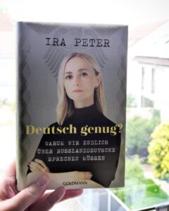Ira Peter - Deutsch genug? Warum wir endlich über
Russlanddeutsche sprechen müssen.
Das Buch wird in einer Wohnung von innen vor ein Fenster gehalten. Im Hintergrund verschwommene Einfamilienhäuser
