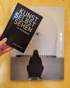 Kunst selbst sehen. Ein Fragenbuch. Eine Hand hält das Buch vor ein Schwarz-Weiß-Foto einer Frau, die eine moderne Skulptur betrachtet.