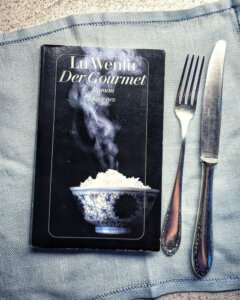 Lu Wenfu, Der Gourmet. Das Taschenbuch liegt auf einer Stoffserviette, daneben Silberbesteck.