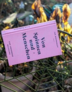 Von Spinnen und Menschen. Das Buch liegt an einem metallenen Spinnennetz in einem herbstlichen Garten