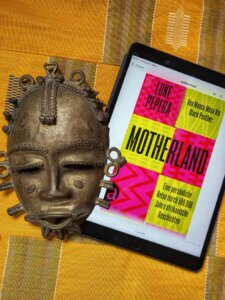 Luke Pepera - Motherland. Das IPad mit dem Cover des E-Books liegt auf einer gleben Tischdecke aus Afrike. Daneben eine Metall-Maske von der Elfenbeinküste.
