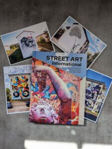 Bildband Street Art International aus dem Midas Verlag liegt auf grauem Boden. Drumherum Postkarten mit Murals von Stadt Wand Kunst in Mannheim