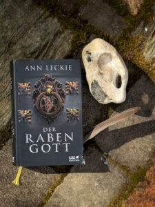 Ann Leckie - Der Rabengott. Das Buch Liegt neuben einer Feder und einem Stein, der wie ein Vogelkopf geformt ist