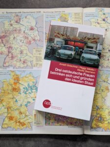 Drei ostdeutsche Frauen betrinken sich und gründen den idealen Staat. Das Buch liegt auf einem alten Schulatlas. Aufgeschlagen sind Karten, die BRD und DDR vergleichen